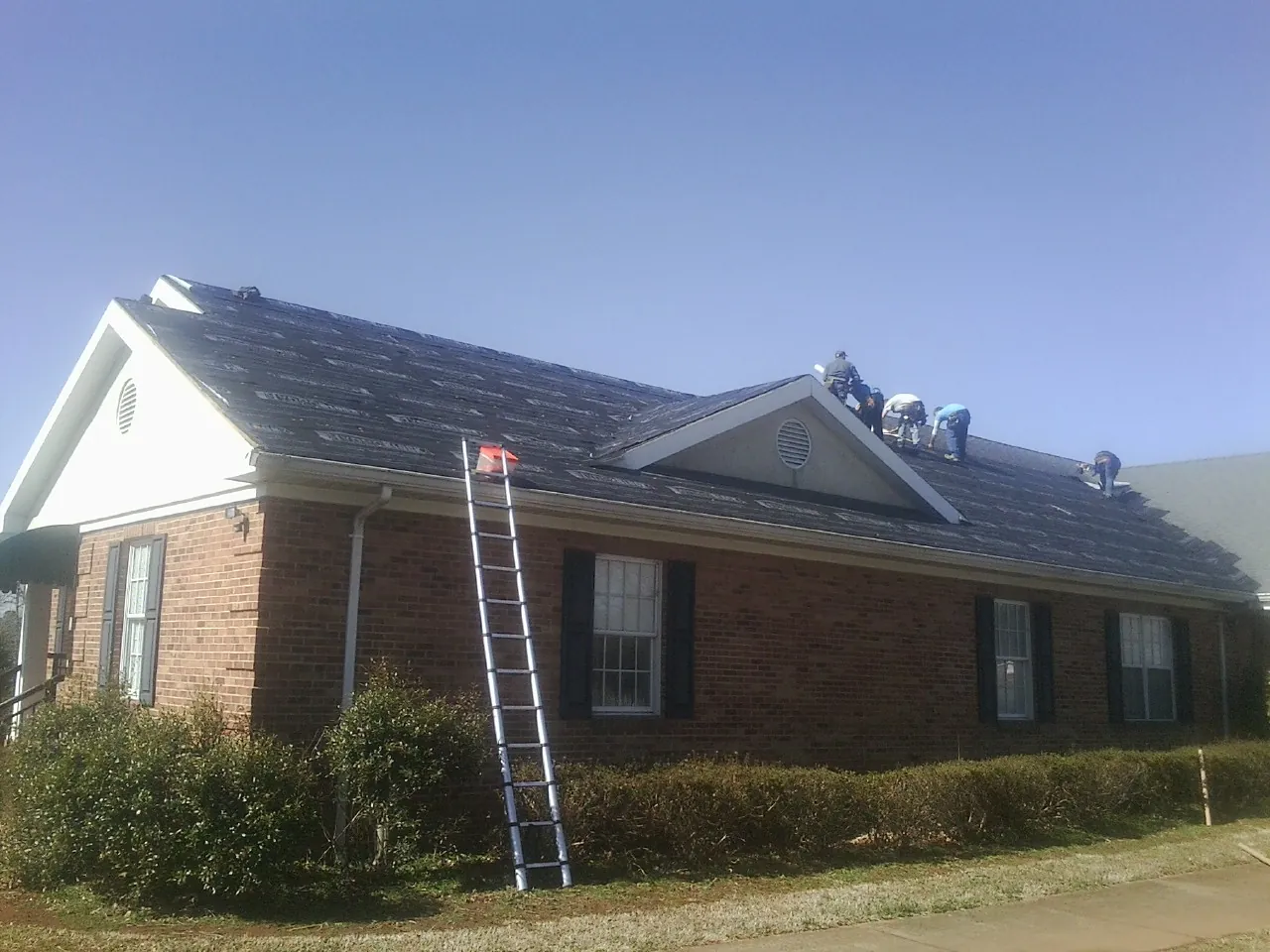 Crew tearing shingles off a brick ranch roof with a ladder set — Emergency Roof Repair work by our crew in Guyton