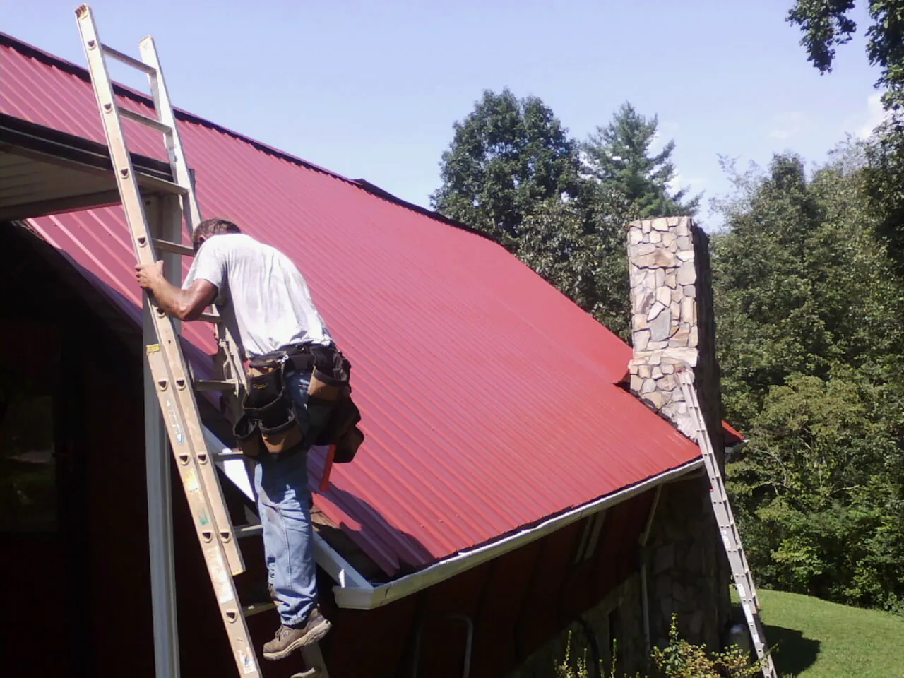 Roofer on a ladder beside a red metal roof and stone chimney — Soffit & Fascia Repair by Roofing Brothers in Guyton